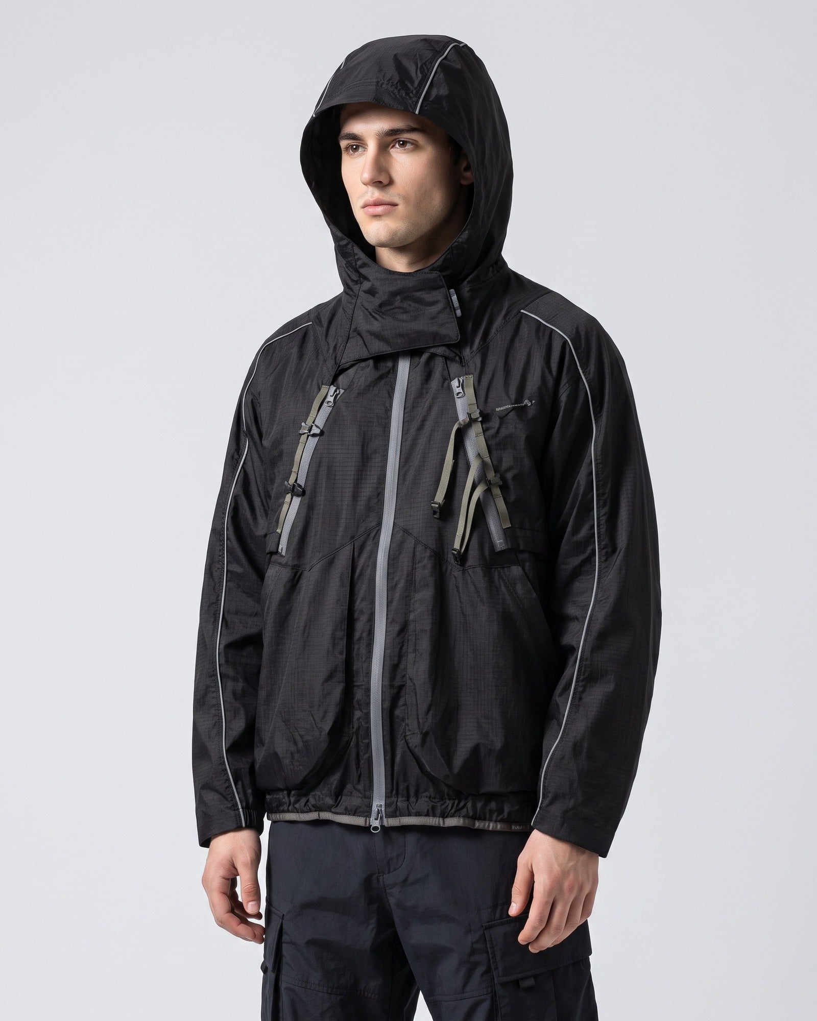 Midnight Black 2-in-1 detachable hooded shell jacket on young European male model, hood up, showing X-webbing chest buckles and reflective piping, clean studio techwear