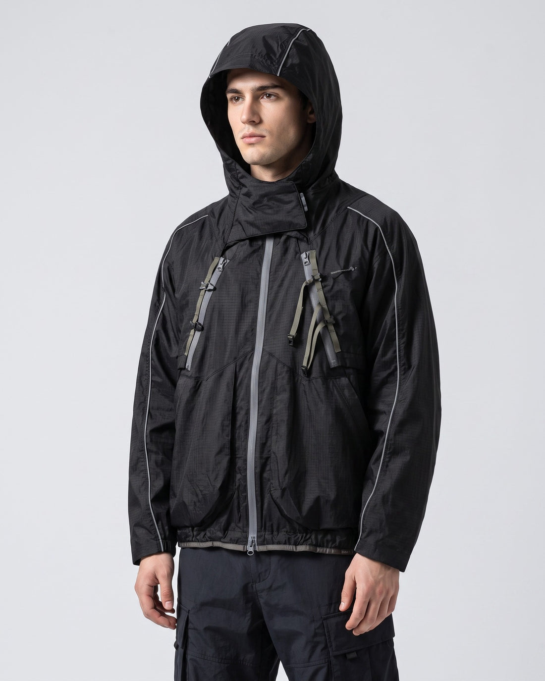 Midnight Black 2-in-1 detachable hooded shell jacket on young European male model, hood up, showing X-webbing chest buckles and reflective piping, clean studio techwear